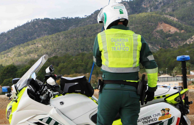 Archivo - Un agente de la Guardia Civil de Tráfico, de espaldas, en una carretera junto a su moto y una montaña al fondo.